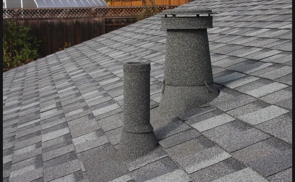 Two stone coated vent stacks on a grey shingle roof beside a backyard fence