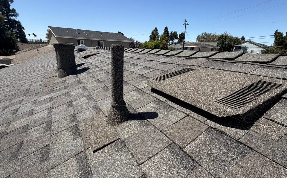 Stone coated pipe and a flat vent on a grey shingle roof under a blue sky