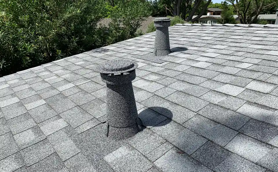Grey stone coated vent with a cap on a platinum shingle roof, trees behind