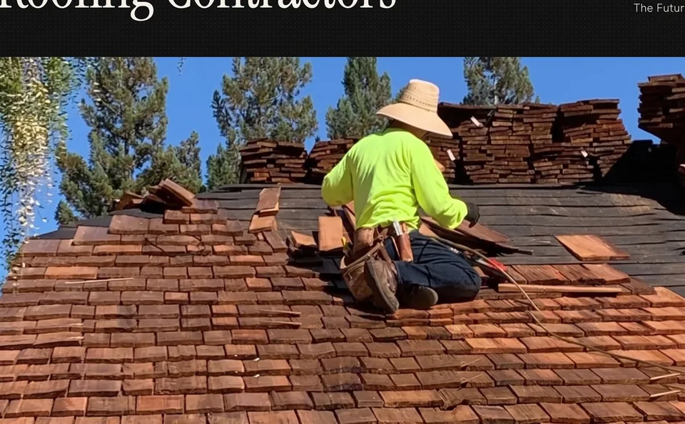 A roofer kneeling on a roof deck installs new cedar shake shingles, stacked shake bundles and pine trees behind him under a clear blue sky